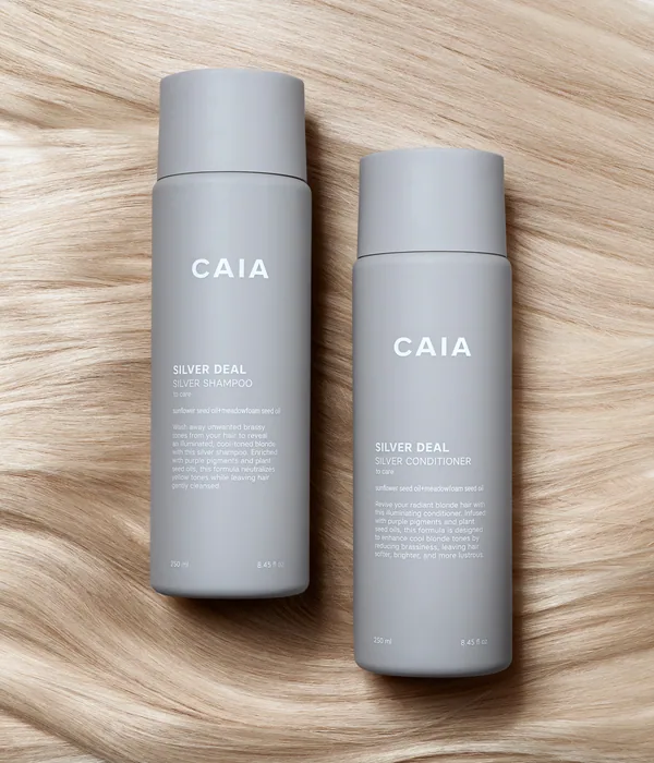 CAIA Silver Deal schampo och balsam i grå flaskor som ligger på blont hår.
